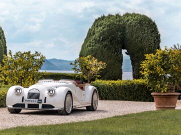 A vintage-style white car is parked on a gravel path, surrounded by green manicured hedges and potted plants, with the beauty of a picturesque water and hilly background adding to the British-Italian atmosphere.