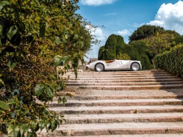 Een Brits-Italiaanse schoonheid, een witte vintage cabriolet, wordt tentoongesteld op een brede stenen trap omgeven door groen en een gedeeltelijk bewolkte hemel, die de elegantie van een Morgan Midsummer belichaamt.