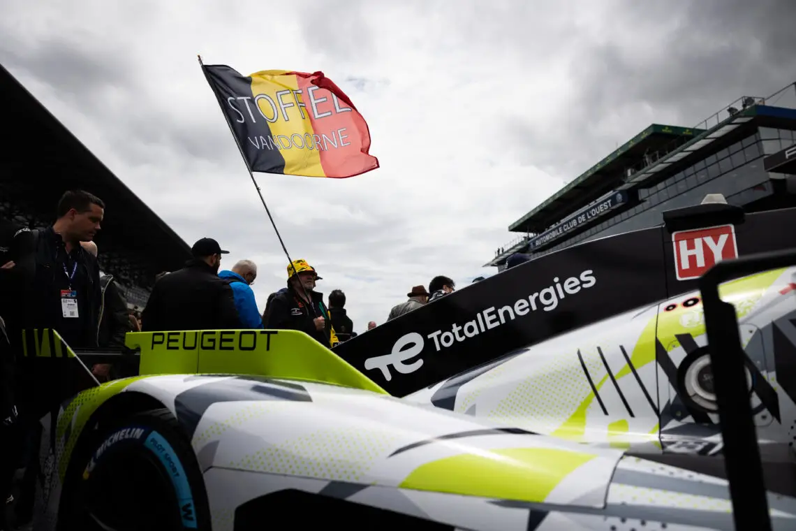 Er wordt een raceauto van Peugeot tentoongesteld, herkenbaar aan de kenmerkende markeringen, met een vlag "Stoffel Vandoorne" op de achtergrond. Enthousiastelingen en racefunctionarissen omringen de Peugeot in een levendige autosportevenementomgeving.