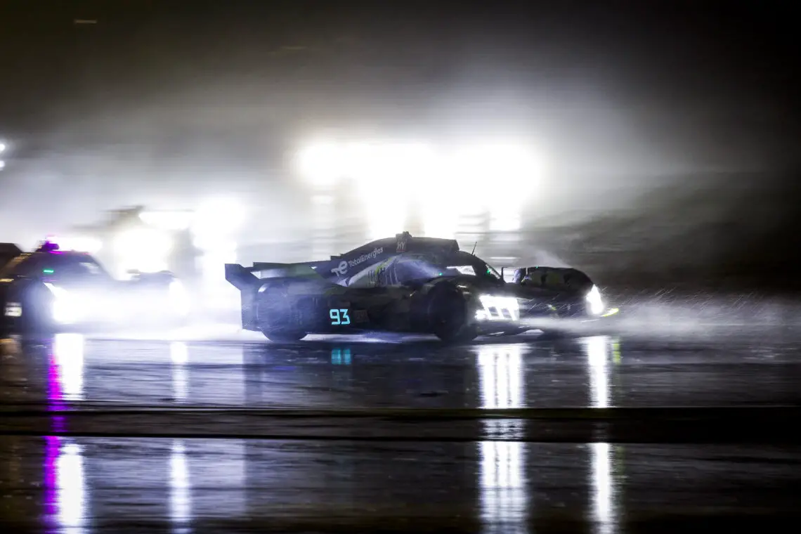 Raceauto nummer 93, een gestroomlijnde Peugeot, snelt 's nachts op een natte baan, terwijl de koplampen reflecteren op het met regen doordrenkte oppervlak. De koplampen van andere voertuigen lijken wazig op de achtergrond.