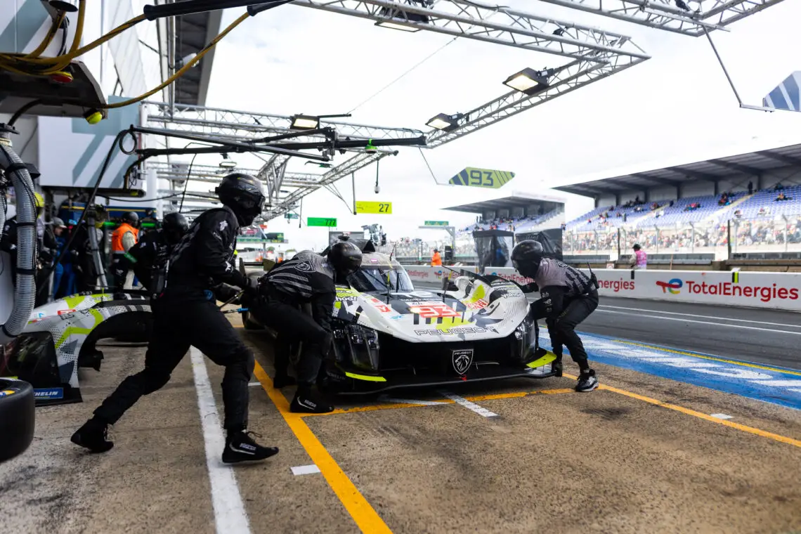 Peugeot-raceauto in pitstop met drie bemanningsleden die onderhoud uitvoeren, omringd door racebaaninfrastructuur en tribunes. De auto is voorzien van prominente sponsorlogo's.