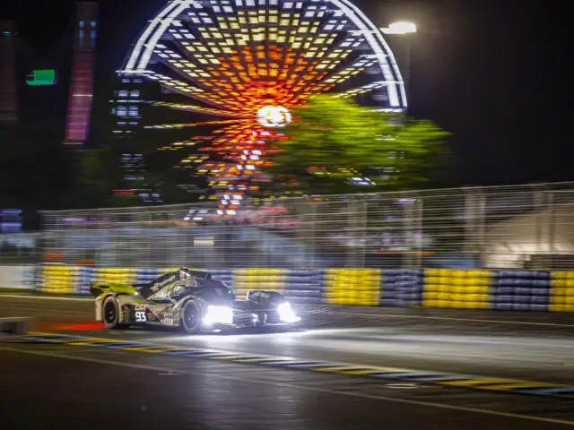 Een raceauto snelt 's nachts over een circuit, verlichte koplampen schijnen, met een verlicht reuzenrad en onscherpe achtergrond in de verte: het is een reportage van de legendarische 24 uur van Le Mans, een ervaring één keer in je leven.