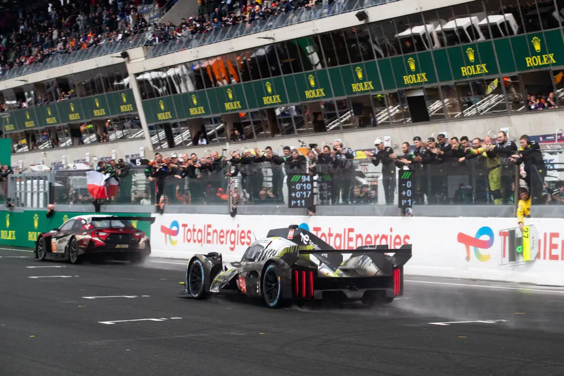 Twee Peugeot-raceauto's snellen langs de finish tijdens een race-evenement, met juichende toeschouwers op de tribunes en verschillende merkbanners zichtbaar.