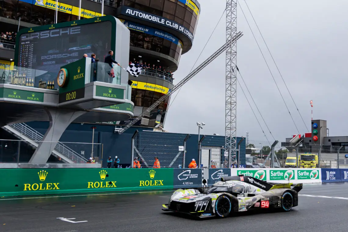 Een Peugeot-sportwagen met het nummer 94 passeert de finishlijn van een racecircuit. Op de achtergrond zijn een timingtoren, toeschouwers en de tribune zichtbaar.