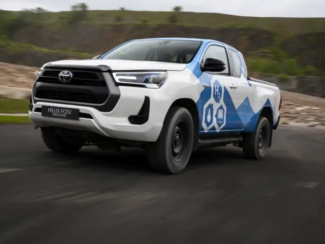 Een witte Toyota Hilux pick-up met een blauw waterstof-waterstofbrandstofcel-ontwerp rijdt op een weg in een landelijk gebied.