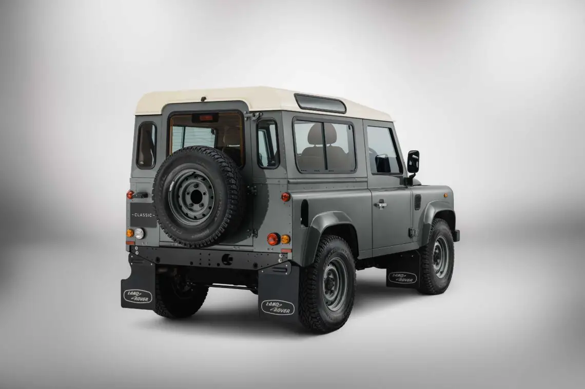 Vue arrière d'un Land Rover Defender gris classique, rappelant l'ère DAKTARI, avec un toit blanc et une roue de secours arrière sur un fond épuré et minimaliste.