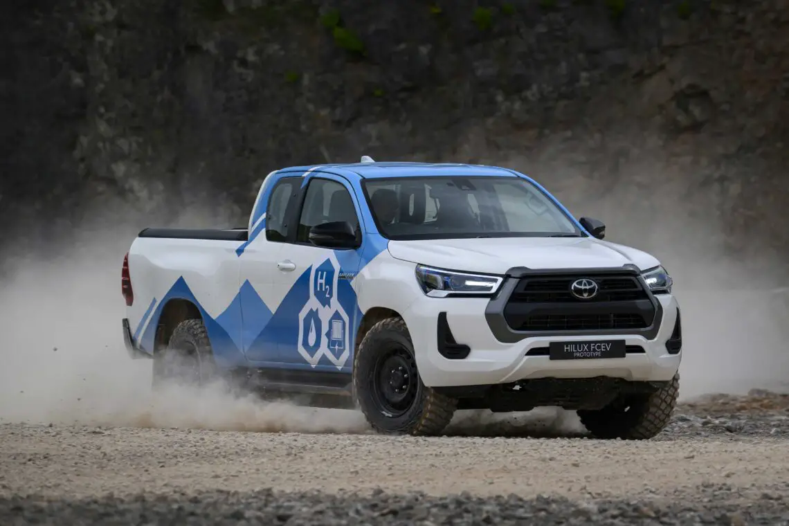 A white-and-blue Toyota Hilux FCV pickup drives down a dirt road, raising dust. The vehicle has an H2O symbol on the side, indicating it is a hydrogen fuel cell vehicle powered by hydrogen technology.
