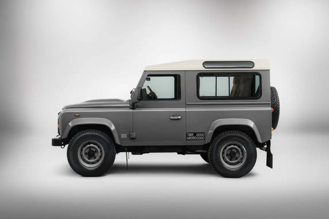 Vue latérale d'un Land Rover Defender gris et blanc avec des pneus épais et une roue de secours à l'arrière, sur un fond blanc uni. Idéal pour votre prochain safari.
