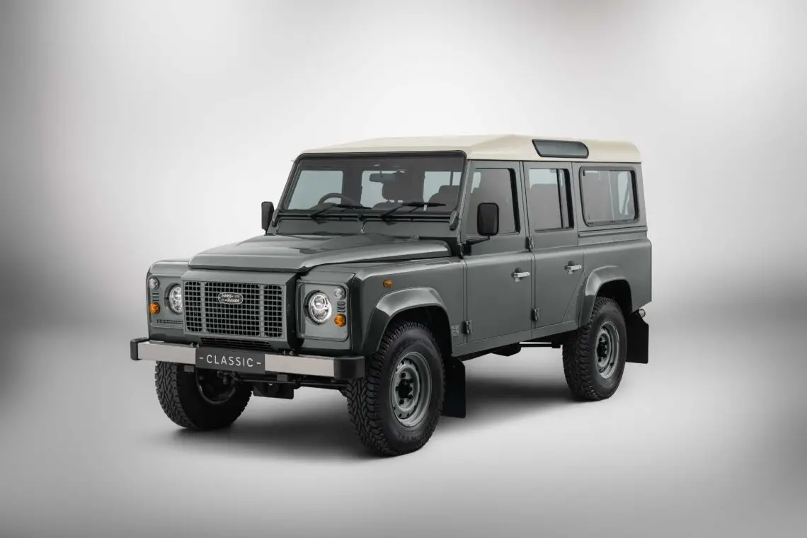 Un Defender au design cube, avec quatre portes et de gros pneus, est exposé dans un studio bien éclairé. Le véhicule a une carrosserie grise et un toit crème, en hommage à l'héritage de Land Rover Classic.