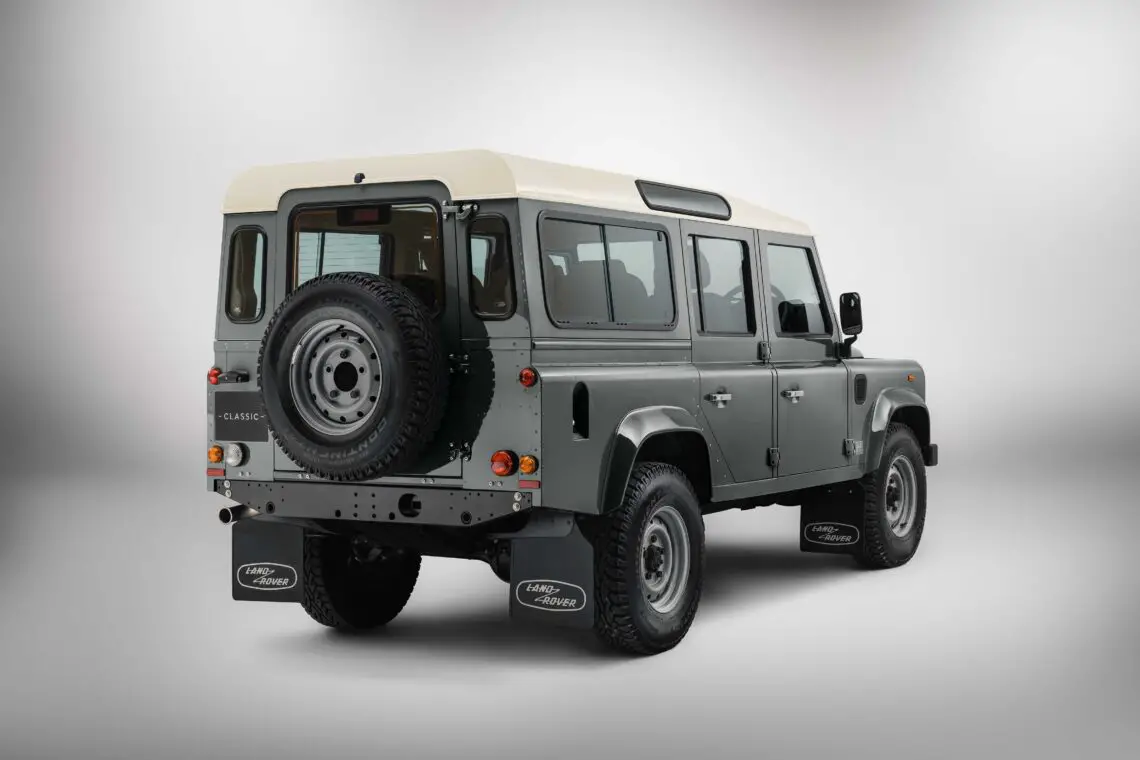 Rear view of a classic green Land Rover Defender 4x4 SUV with a white roof, black mud flaps and a spare tire on the back, reminiscent of a rugged safari adventure, positioned against a plain gray background.
