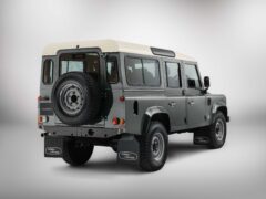 Achteraanzicht van een grijze Land Rover Defender Classic. Het voertuig, dat doet denken aan het DAKTARI-tijdperk, heeft een wit dak, een reservewiel achterop en spatlappen met het Land Rover-logo. Het wordt tentoongesteld in een studiosetting.