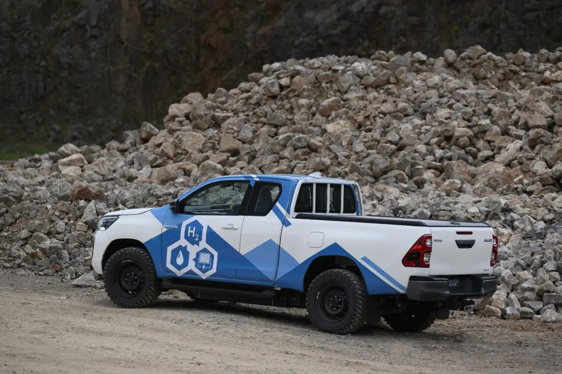 Una camioneta Toyota impulsada por hidrógeno, de color blanco y azul, marcada como hydrogen-Hilux, está aparcada cerca de un montón de piedras sobre una superficie de grava. El vehículo presenta diseños y símbolos que indican el sistema de combustible de hidrógeno.