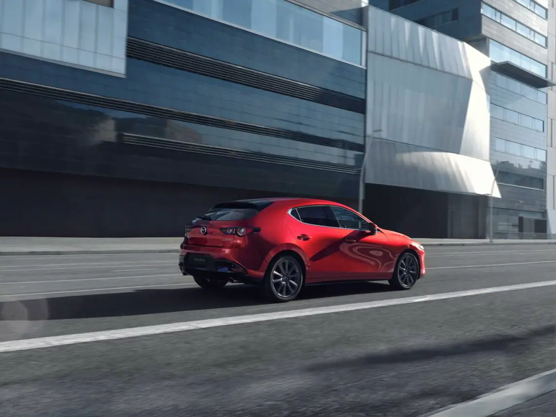 Een rode Mazda3 hatchback, modeljaar 2025, rijdt door een lege stadsstraat met moderne glazen gebouwen op de achtergrond.