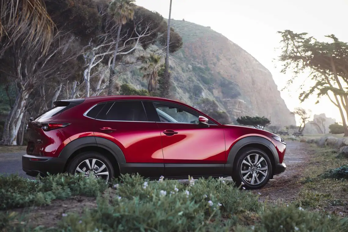 Een rode Mazda CX-30 staat geparkeerd op een schilderachtige, ruige kustweg met heuvels, bomen en de oceaan op de achtergrond.
