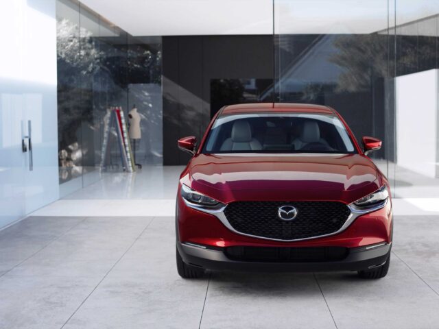 Vooraanzicht van een rode Mazda3 CX-30 geparkeerd in een modern gebouw met glazen wanden met lichtreflecties op het oppervlak, wat het elegante ontwerp van modeljaar 2025 laat zien.