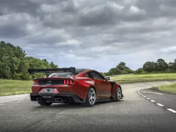 A red Ford Mustang GTD with rear spoiler is parked on a winding road surrounded by greenery and under a cloudy sky.
