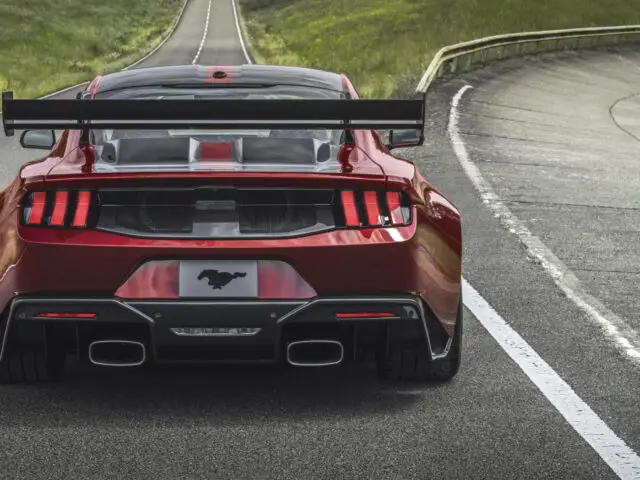 A red Ford Mustang GTD with a large rear spoiler and dual exhaust pipes drives on a winding road through a grassy landscape, showing off its skill and prize as it maneuvers effortlessly.