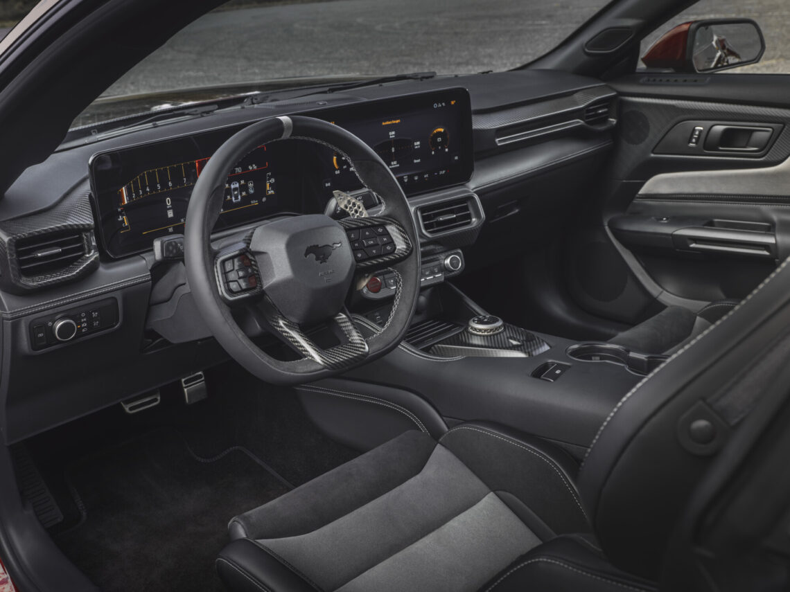 Innenansicht eines modernen Ford Mustang GTD mit Lenkrad, digitalem Armaturenbrett und verschiedenen Bedienelementen. Das Design ist in Schwarz- und Grautönen mit Akzenten aus Karbonfaser gehalten.