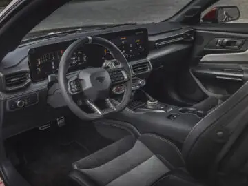 Vue de l'intérieur d'une Ford Mustang GTD moderne avec volant, tableau de bord numérique et diverses commandes. Le design présente des tons noirs et gris avec des accents de fibre de carbone.