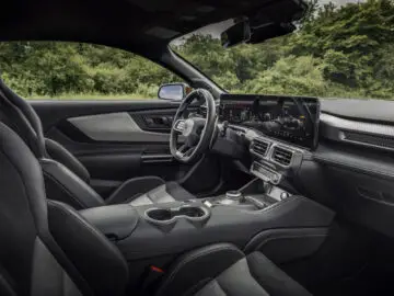 Interior de un Ford Mustang GTD moderno con asientos de cuero negro, un salpicadero detallado con una gran pantalla y un elegante volante. La vegetación es visible a través de las ventanillas.