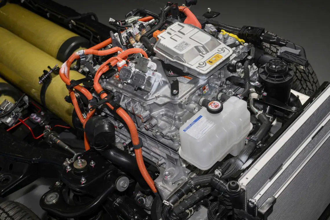 Close-up of the exposed motor and battery components of an electric vehicle, showing various cables, connectors and reservoirs in a mechanical arrangement. This detailed image highlights the innovative engineering behind the Toyota Hilux's advanced powertrain.