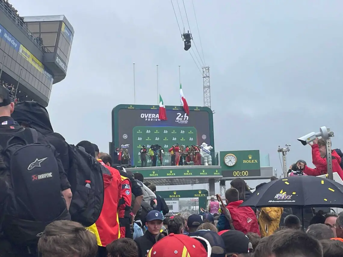 Menigte kijkt naar coureurs op het podium op een racecircuit tijdens een prijsuitreiking. Drie coureurs staan op verschillende niveaus van het podium met vlaggen en schermen die de resultaten op de achtergrond weergeven, waardoor een reportagewaardig moment wordt vastgelegd. Dit is werkelijk een ‘één keer in je leven’-ervaring, die doet denken aan de 24 uur van Le Mans.