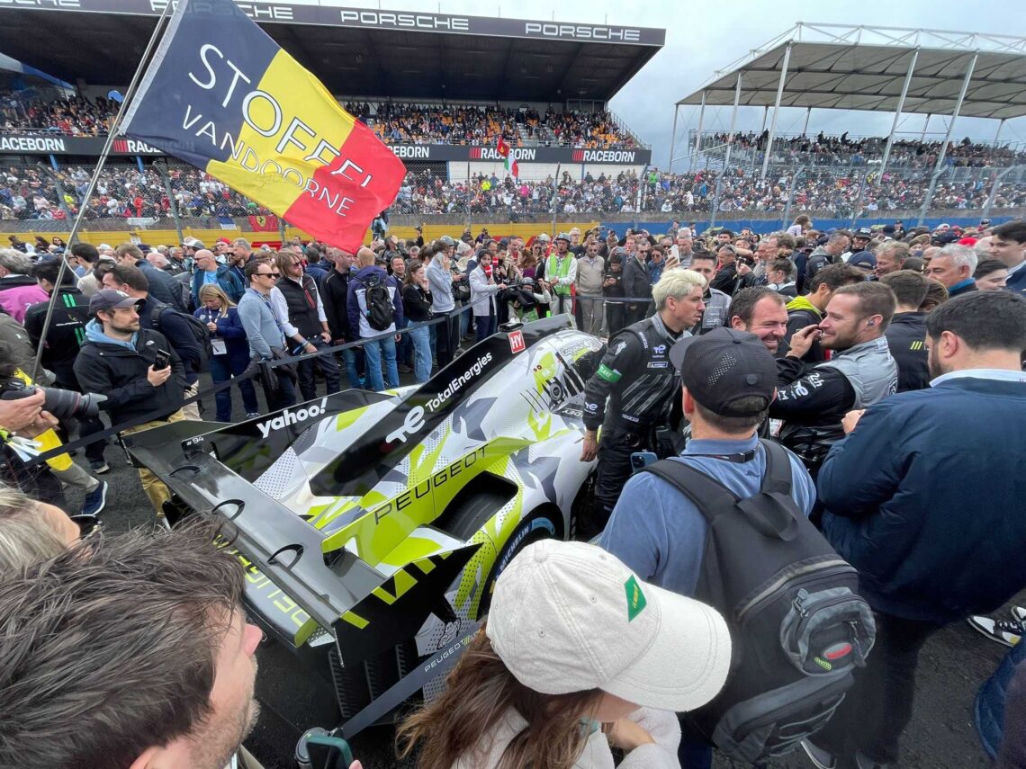 Massa's mensen omsingelen een Peugeot-raceauto op het circuit van de 24 uur van Le Mans, terwijl er met de vlag "Stoffel Vandoorne" wordt gezwaaid. Onder een bewolkte hemel zijn op de achtergrond tribunes vol toeschouwers zichtbaar, die het levendige leven van dit iconische autorace-evenement vastleggen.