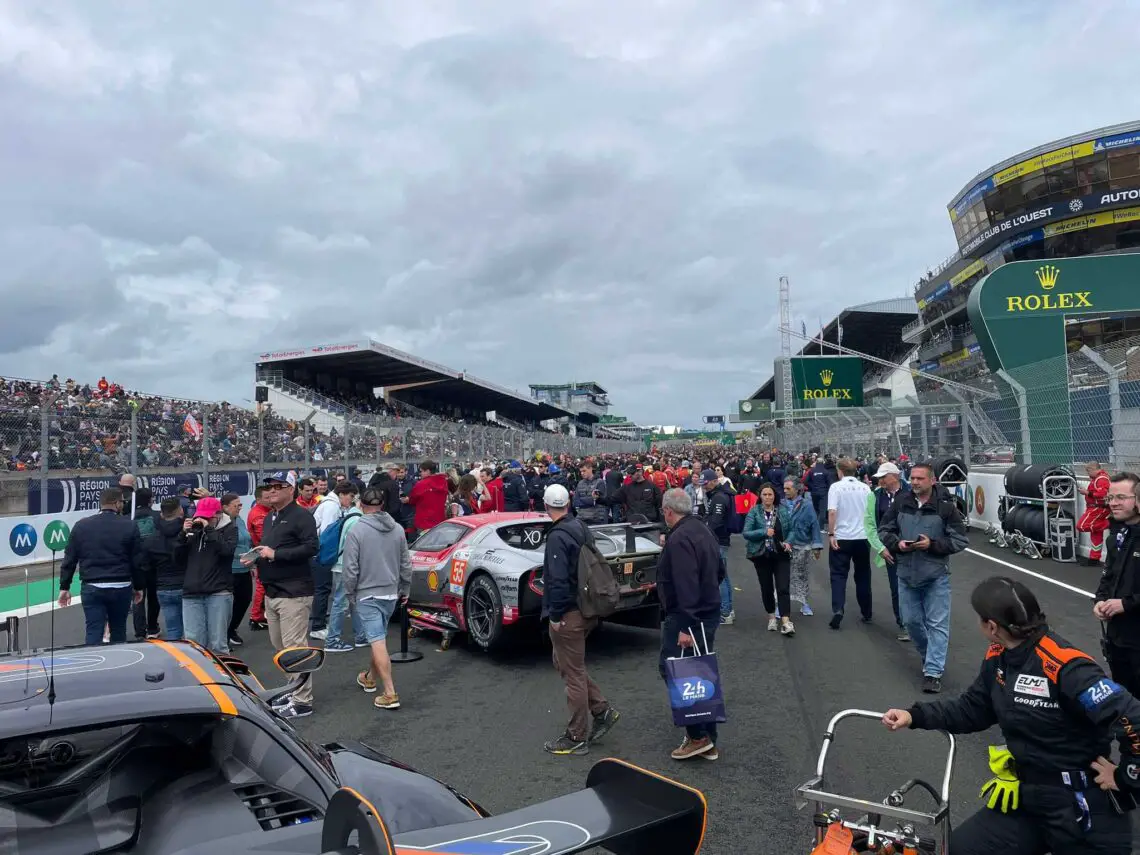 Een grote menigte verzamelt zich op het circuit en geniet van de opwinding van de 24 uur van Le Mans terwijl auto's en teampersoneel zich voorbereiden op dit unieke evenement; De tribunes en Rolex-borden zijn zichtbaar op de achtergrond en vormen een perfecte scène voor reportages.