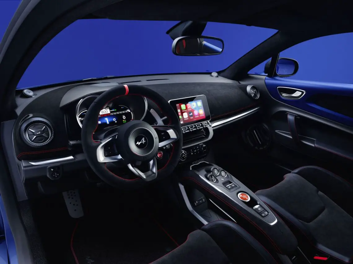 De afbeelding toont het interieur van een moderne Alpine-sportwagen met zwarte en rode accenten, voorzien van een multifunctioneel stuur, digitaal display, touchscreen-infotainmentsysteem en verschillende bedieningsknoppen. Dit A110-model toont de toewijding van het merk aan precisie en stijl via zijn personalisatieprogramma.