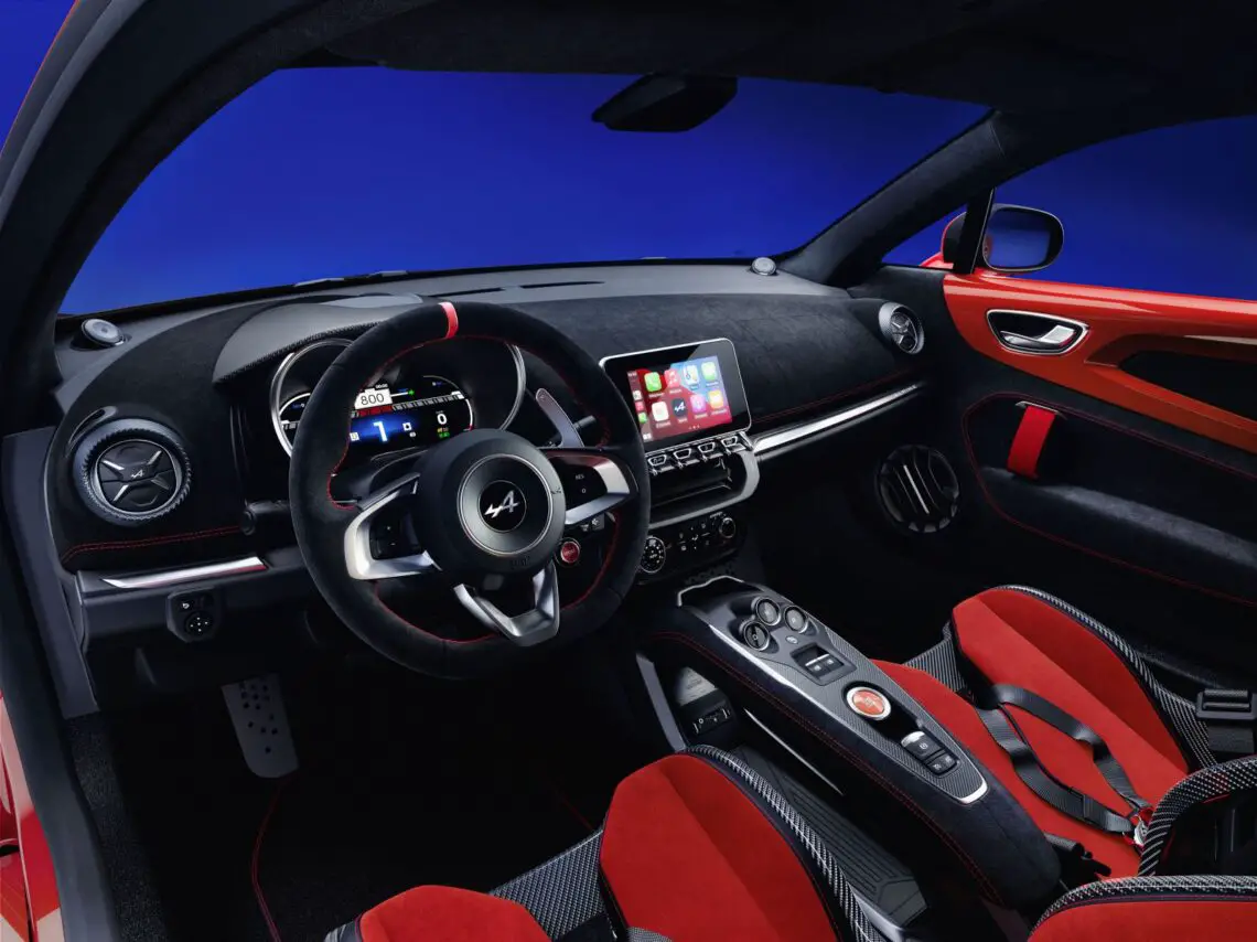 Interieur van een Alpine A110-sportwagen met een stuur met logo, digitaal dashboard, touchscreendisplay, bedieningsknoppen en stoelen met rode accenten en zwarte bekleding. Verbeter uw rit met ons personalisatieprogramma voor een unieke rijervaring op maat.