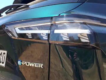 Close-up van het achterlicht en het e-POWER-embleem op een donkergekleurde Nissan Qashqai. Het ontwerp is voorzien van strakke, hoekige lijnen met reflecties van de omgeving die zichtbaar zijn op het oppervlak van de auto, wat een kijkje geeft in de hoogtepunten van de review van 2024.