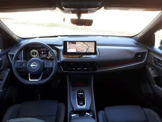 Interieur van een Nissan Qashqai met het strakke dashboard en het stuurwiel versierd met het Nissan-logo, een touchscreen-infotainmentsysteem met navigatie, bedieningsknoppen en andere moderne designelementen, zoals benadrukt in de recensie van 2024.