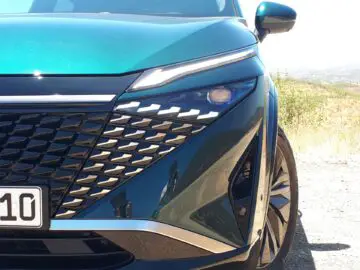 Close-up van de rechtervoorzijde van een blauwgroen Nissan Qashqai met een futuristisch ontwerp, waarbij de koplamp, de grille en een deel van de kentekenplaat te zien zijn terwijl het bergachtige landschap wordt weerspiegeld op het glanzende oppervlak van de auto.
