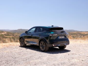 Een zwarte Nissan Qashqai staat geparkeerd op een grindgebied met uitzicht op een schilderachtig, bergachtig landschap onder een heldere, blauwe lucht.