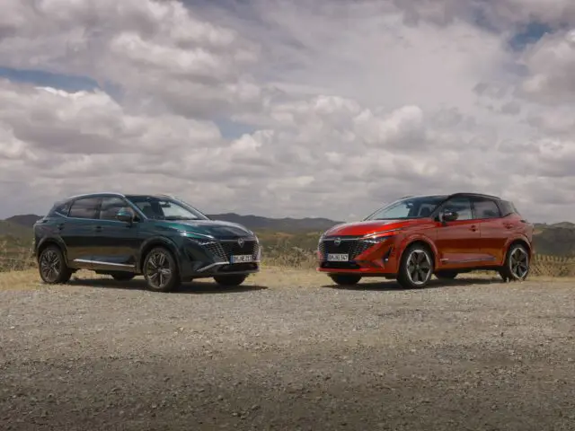 Twee geparkeerde SUV's, een groene en een rode, staan op een onverharde weg tegen de achtergrond van glooiende heuvels en een bewolkte lucht. Deze scène zou rechtstreeks uit een recensie uit 2024 kunnen komen voor de stoere maar stijlvolle Nissan Qashqai.