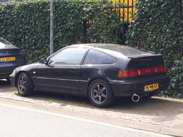 Une Honda CRX noire d'origine est garée dans une rue à côté d'une clôture verte envahie par le lierre aux Pays-Bas. Repérée dans un état impeccable.