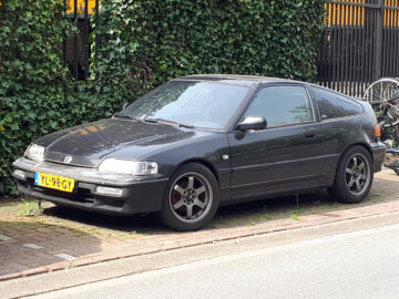 Een zwarte tweedeurs Honda CR-X staat geparkeerd aan de kant van een straat, naast een met groene klimop bedekt hek. Er staan fietsen achter de auto. Op het Nederlandse kenteken staat "YL-939-GY.