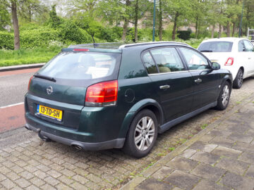 A green Opel Signum hatchback is parked on the street. Spotted with European registration number 13-TD-SH, in front of the vehicle is partially visible a white car. Trees and greenery adorn the background.