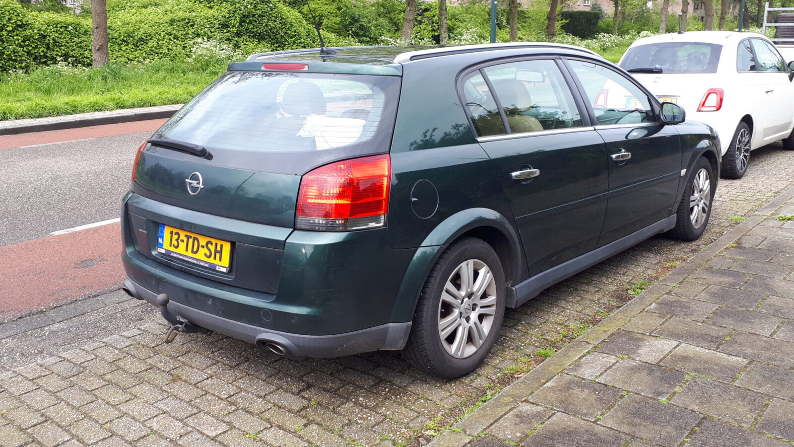 In de straat staat een donkergroene Opel-stationwagen, mogelijk een Gespot Signum, naast een witte auto geparkeerd. De auto is voorzien van een trekhaak en een Nederlands kenteken met de tekst "13-TD-SH". Op de achtergrond zijn bomen en groen zichtbaar.