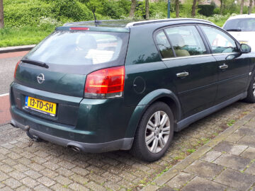 In de straat staat een donkergroene Opel-stationwagen, mogelijk een Gespot Signum, naast een witte auto geparkeerd. De auto is voorzien van een trekhaak en een Nederlands kenteken met de tekst "13-TD-SH". Op de achtergrond zijn bomen en groen zichtbaar.