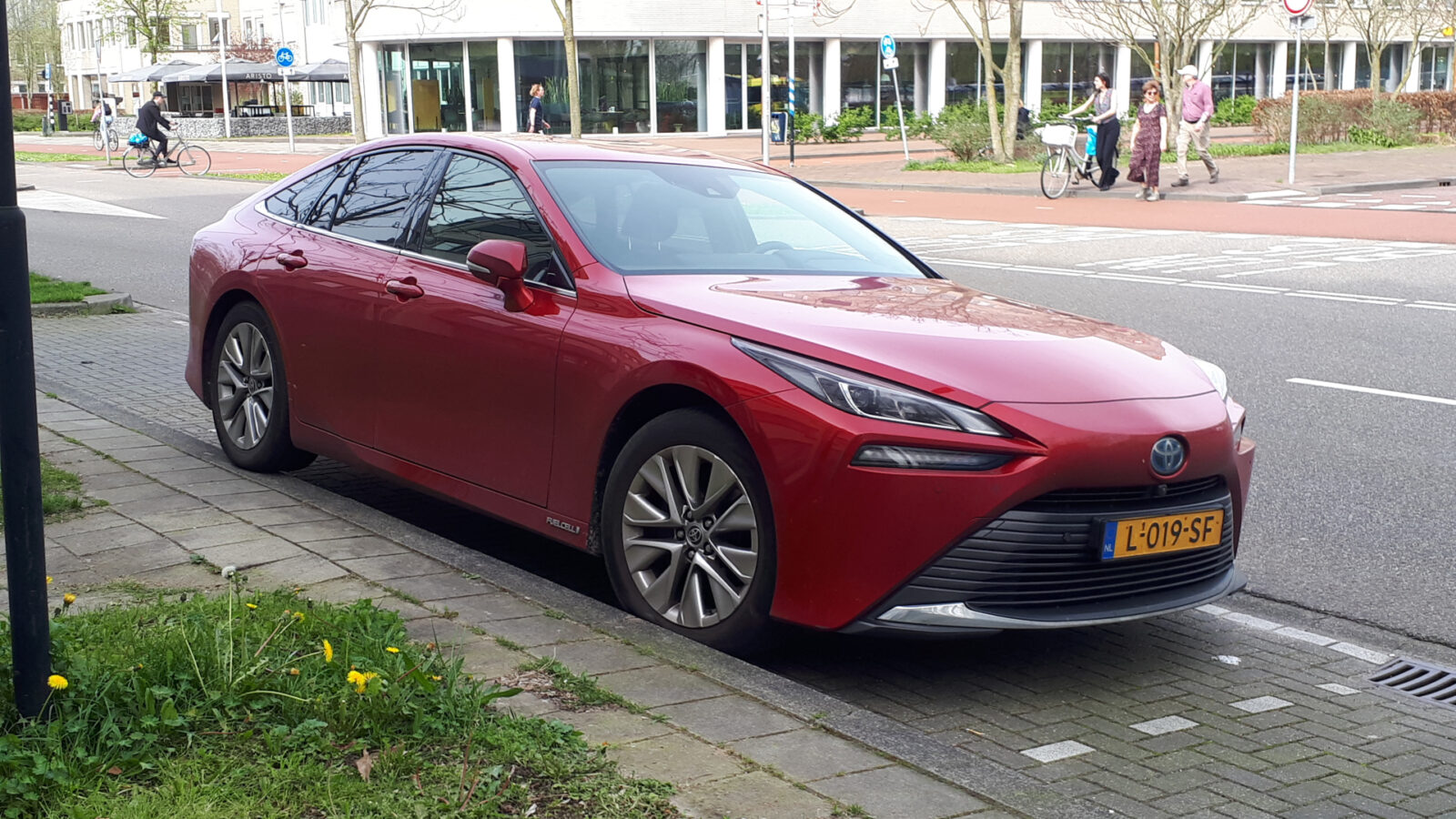 Een rode Toyota Mirai sedan met kenteken L-019-SF staat Gespot parallel aan de stoeprand geparkeerd in een straat met fietspaden en voetgangers op de achtergrond.