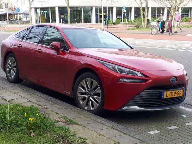 Een rode Toyota Mirai sedan met kenteken L-019-SF staat Gespot parallel aan de stoeprand geparkeerd in een straat met fietspaden en voetgangers op de achtergrond.