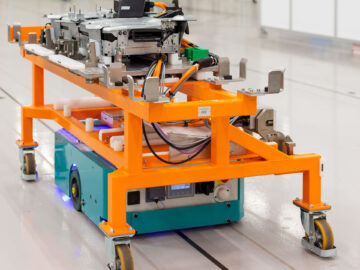 Een automatisch geleid voertuig transporteert een mechanisch samenstel op een oranje frame door een schone, industriële omgeving, die doet denken aan de precisie die te vinden is in de Nissan Qashqai-productielijn van Sunderland.