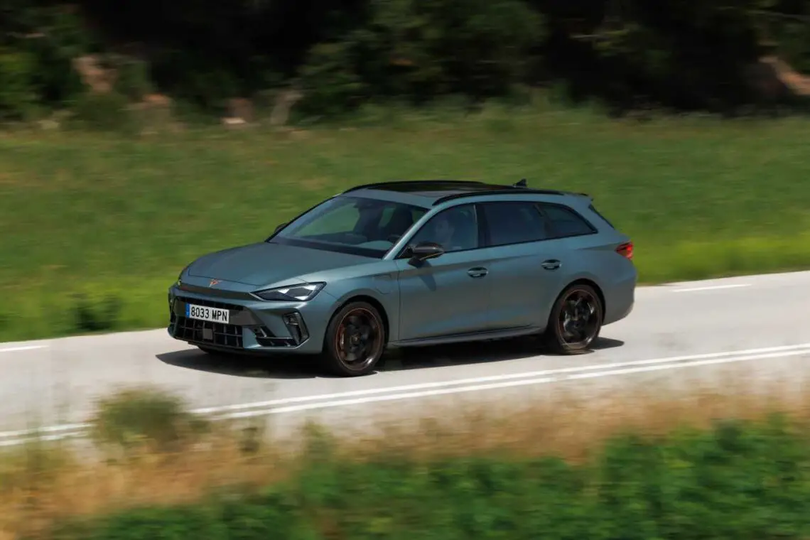 Een strakke grijze stationwagen rijdt over een landelijke weg met groene velden en bomen op de achtergrond, die doet denken aan een stijlvolle Cupra Leon in alle rust.