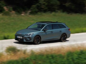 Een strakke grijze stationwagen rijdt over een landelijke weg met groene velden en bomen op de achtergrond, die doet denken aan een stijlvolle Cupra Leon in alle rust.
