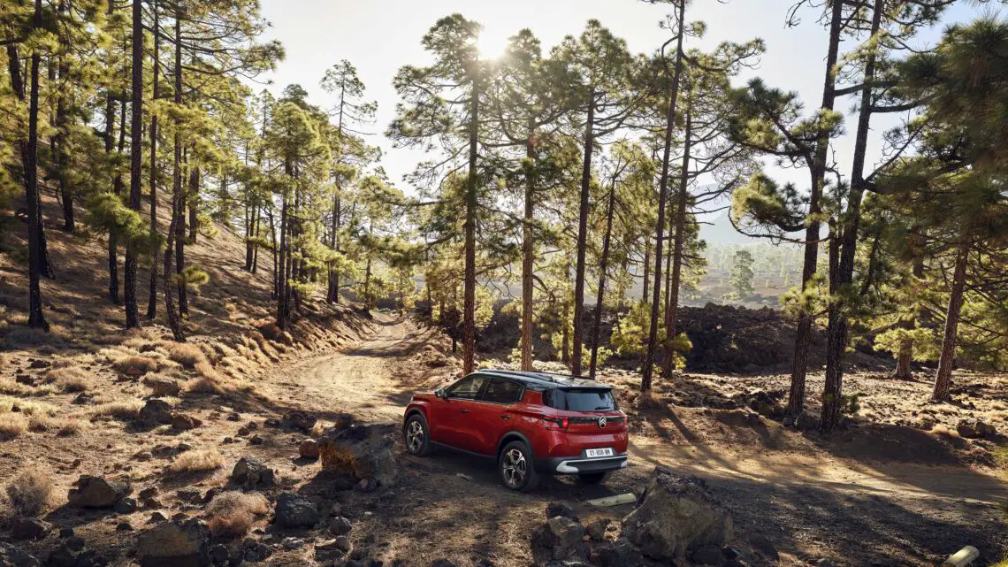 Een Citroën (ë)-C3 Aircross, geprijsd op 25.850 euro, staat geparkeerd op een onverharde weg in een zonovergoten bos met hoge dennenbomen en rotsachtig terrein.