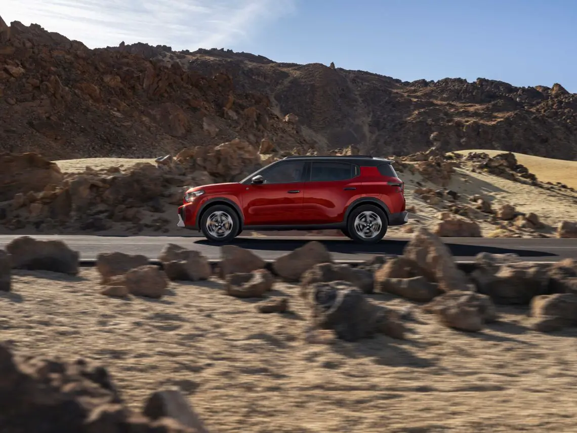Een rode Citroën (ë)-C3 Aircross SUV rijdt over een weg door een rotsachtig, woestijnlandschap met bergen op de achtergrond onder een heldere hemel. Met een prijskaartje van 25.850 euro combineert deze strakke zevenzitter moeiteloos stijl met avontuur.