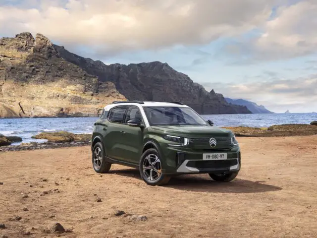 Een groen-witte Citroën C3 Aircross SUV staat geparkeerd op een zandig kustgebied met ruige rotswanden en de oceaan op de achtergrond.