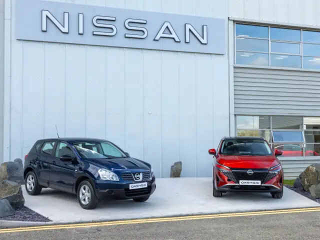 Twee Nissan Qashqai-voertuigen worden buiten een Nissan-gebouw tentoongesteld; de ene is blauw en de andere is rood, beide geparkeerd op een betonnen ondergrond met een groot Nissan-bord erboven, waarop de nieuwste modellen van het merk worden getoond.