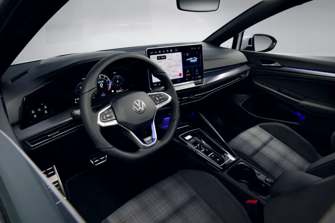 Het interieur van de voorstoelen van de Volkswagen Golf EV omvat een stuur met VW-logo, een digitaal dashboard, een centraal touchscreen met navigatie- en rijbereik en diverse bedieningselementen.